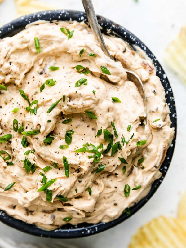 A close up shot of caramelized onion dip inside of a blue and white speckled bowl for serving. The onion dip is garnished with freshly snipped chives and ground black pepper. The bowl sits atop a creamy white textured surface with a gray and white linen napkin and loose ruffle potato chips surrounding the bowl of dip at center. A silver spoon rests inside of the bowl of dip for serving.