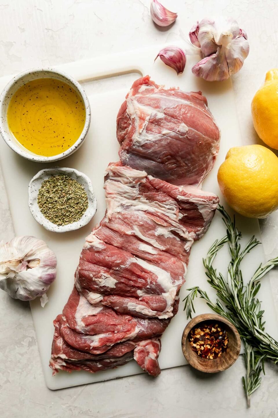 Butterflied grilled leg of lamb ingredients arranged on a creamy white textured surface: boneless leg of lamb, olive oil, lemons, garlic, fresh rosemary, dried oregano, crushed red pepper flakes, kosher salt, and ground black pepper.