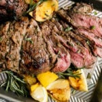 A partially carved grilled leg of lamb rests atop an aluminum baking sheet surrounded by grilled lemons and sprigs of rosemary. The baking sheet pan sits atop a creamy white textured surface with a blue and white striped linen napkin and a small wooden pinch bowl filled with kosher salt resting alongside.