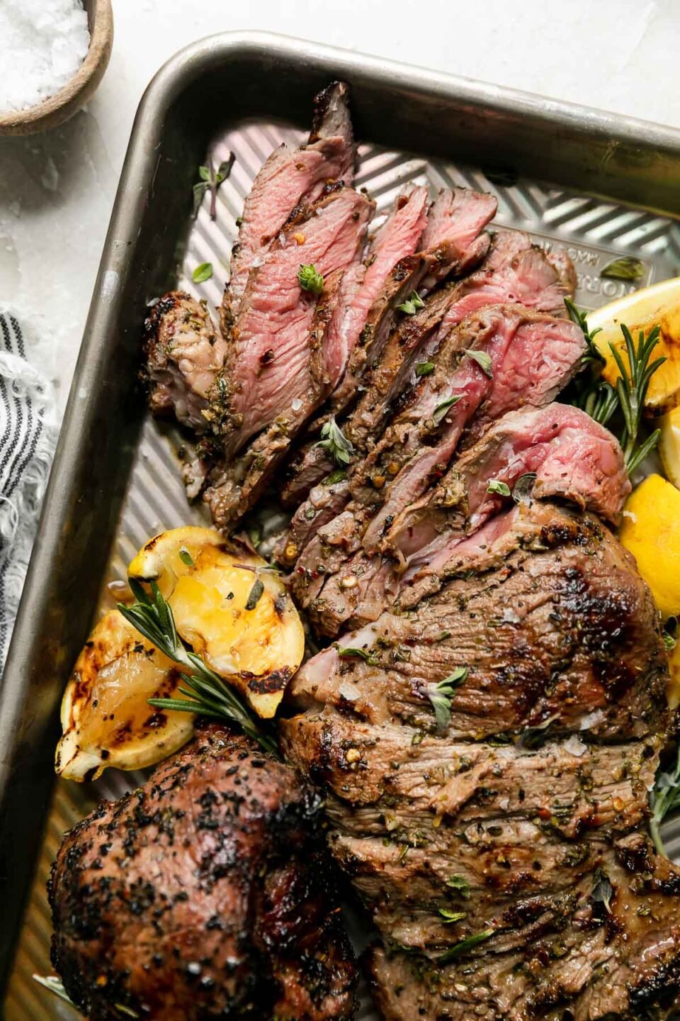 A partially carved grilled leg of lamb rests atop an aluminum baking sheet surrounded by grilled lemons and sprigs of rosemary. The baking sheet pan sits atop a creamy white textured surface with a blue and white striped linen napkin and a small wooden pinch bowl filled with kosher salt resting alongside.