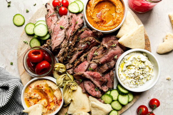 A mezze-style platter served on a round wooden board is filled with thinly sliced grilled leg of lamb, sliced cucumbers, tomatoes, roasted tomatoes, artichoke hearts, dips & spreads, and pita bread. The platter is surrounded by a jar of pickled red onion, cherry tomatoes on the vine, loose fresh herbs, and a blue and white striped linen napkin.