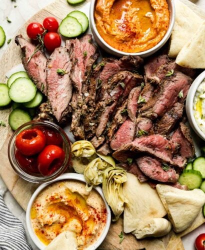 A mezze-style platter served on a round wooden board is filled with thinly sliced grilled leg of lamb, sliced cucumbers, tomatoes, roasted tomatoes, artichoke hearts, dips & spreads, and pita bread. The platter is surrounded by a jar of pickled red onion, cherry tomatoes on the vine, loose fresh herbs, and a blue and white striped linen napkin.