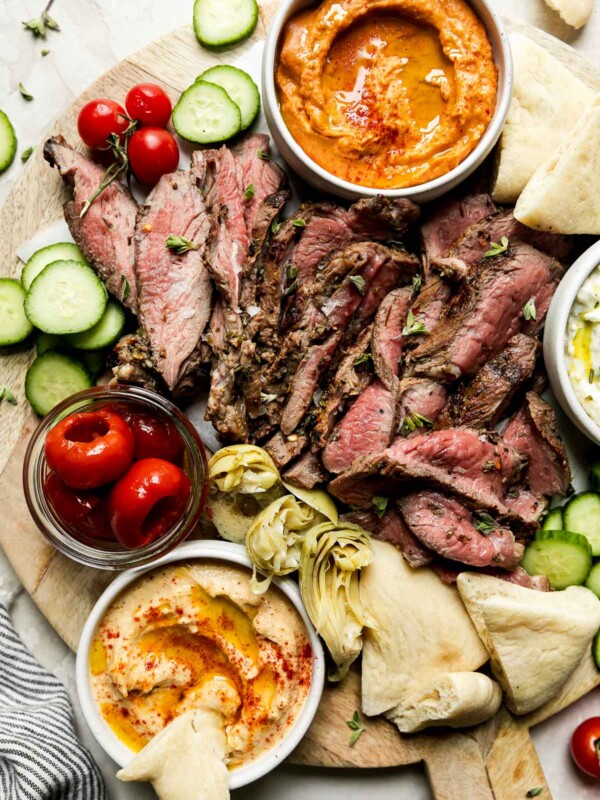 A mezze-style platter served on a round wooden board is filled with thinly sliced grilled leg of lamb, sliced cucumbers, tomatoes, roasted tomatoes, artichoke hearts, dips & spreads, and pita bread. The platter is surrounded by a jar of pickled red onion, cherry tomatoes on the vine, loose fresh herbs, and a blue and white striped linen napkin.