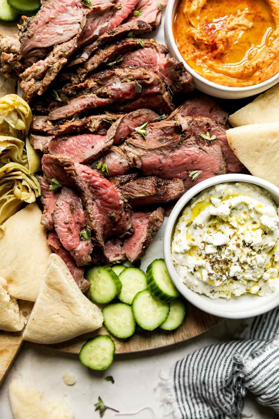 A close up of a mezze-style platter served on a round wooden board topped with thinly sliced grilled leg of lamb, sliced cucumbers, artichoke hearts, dips & spreads, and pita bread. The platter rests atop a creamy white textured surface and a blue and white striped linen napkin rests alongside.