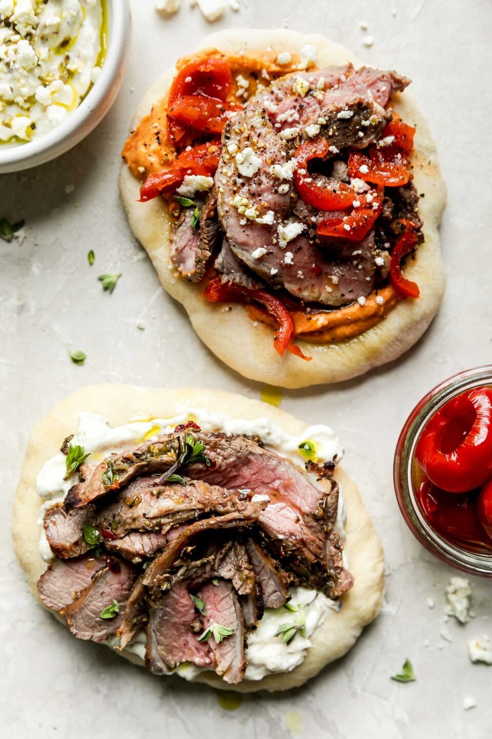 Two small pieces of pita bread are topped with hummus and a Greek yogurt dipped then topped with thinly sliced grilled boneless leg of lamb and other thinly sliced veggies. The two pitas sit atop a creamy white textured surface alongside a small glass jar filled with roasted red peppers and a small white ceramic bowl filled with a Greek-yogurt based dip.