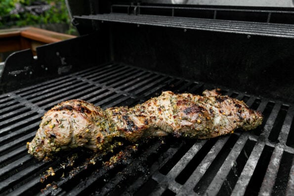 Grilled boneless leg of lamb rests atop gas grill grates. The lamb has been marinated in a lemon and garlic marinade.