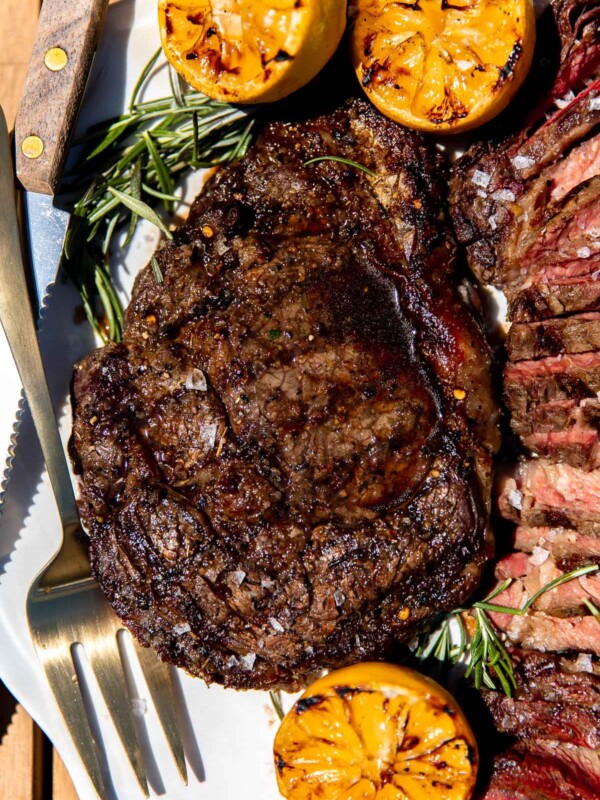 A single grilled whole Tuscan steak rests atop a white serving platter surround by char-grilled lemons, fresh rosemary, thinly sliced Tuscan steak, a steak knife, and a gold serving fork. The platter sits atop a wood dining table.