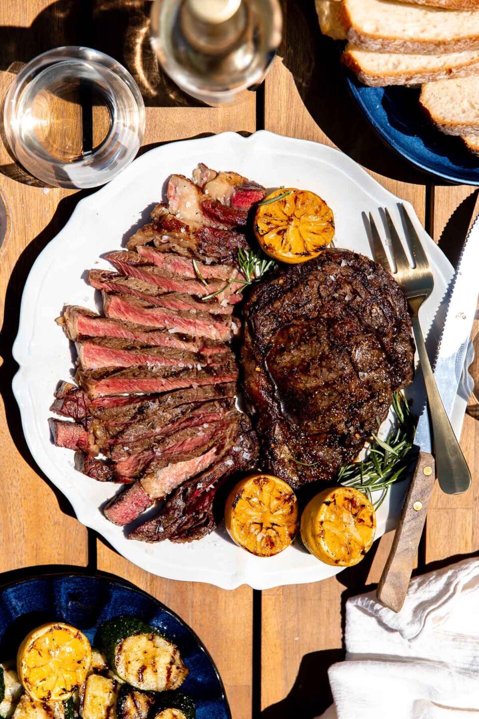 A single grilled whole Tuscan steak rests atop a white serving platter surround by char-grilled lemons, fresh rosemary, thinly sliced Tuscan steak, a steak knife, and a gold serving fork. The platter sits atop a wood dining table and is surrounded by a bottle of white wine, a glass of white wine, a white linen napkin, and two dark blue ceramic plates filled with char-grilled lemons and bread respectively.
