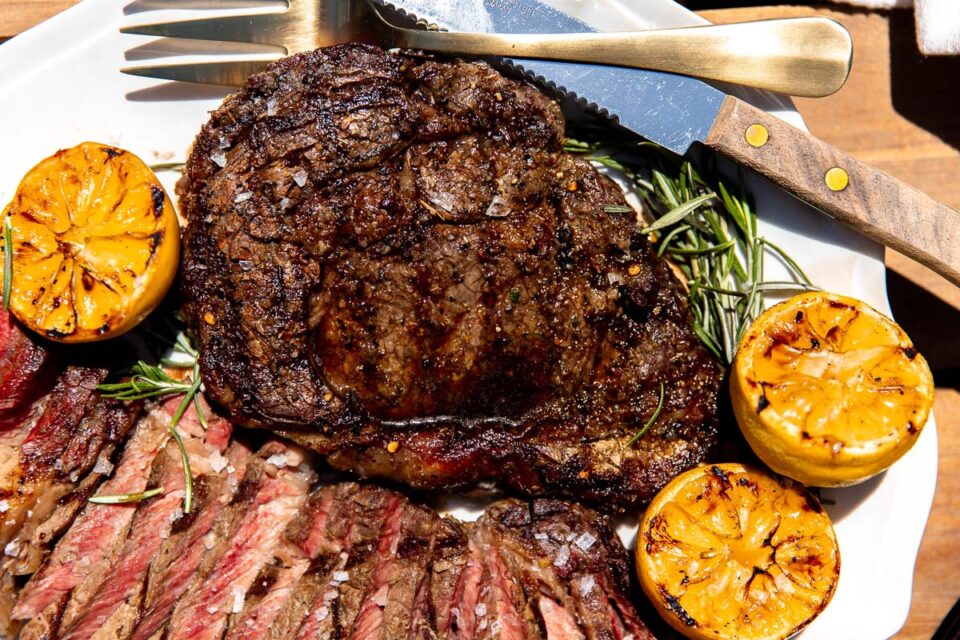 A side angle shot of a single grilled whole Tuscan steak resting atop a white serving platter surround by char-grilled lemons, fresh rosemary, thinly sliced Tuscan steak, a steak knife, and a gold serving fork. The platter sits atop a wood dining table.