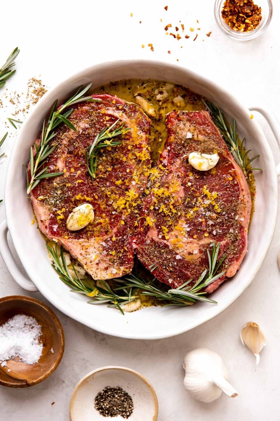 How to make Tuscan style steak: two steaks marinate in a Tuscan-style marinade made with olive oil, lemon zest, garlic, fresh rosemary, dried oregano, and crushed red pepper flakes. The steaks marinate inside of a double handle baking dish that sits atop a creamy white textured surface surrounded by a bulb of garlic and three small pinch bowls filled with crushed red pepper flakes, kosher salt, and ground black pepper.