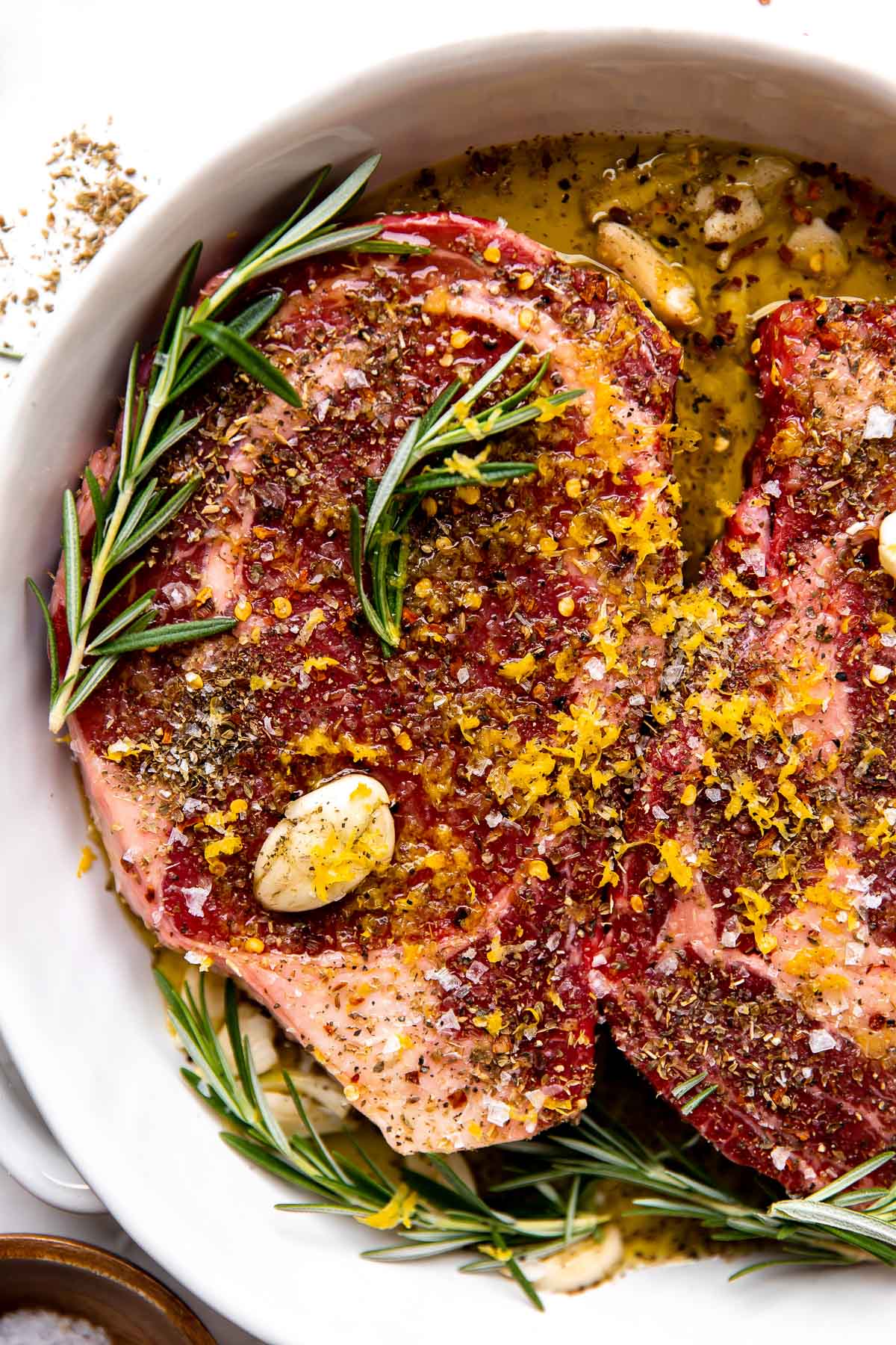 A close up shot of how to make Tuscan style steak: two steaks marinate in a Tuscan-style marinade made with olive oil, lemon zest, garlic, fresh rosemary, dried oregano, and crushed red pepper flakes. The steaks marinate inside of a double handle baking dish that sits atop a creamy white textured surface surrounded by a bulb of garlic and three small pinch bowls filled with crushed red pepper flakes, kosher salt, and ground black pepper.