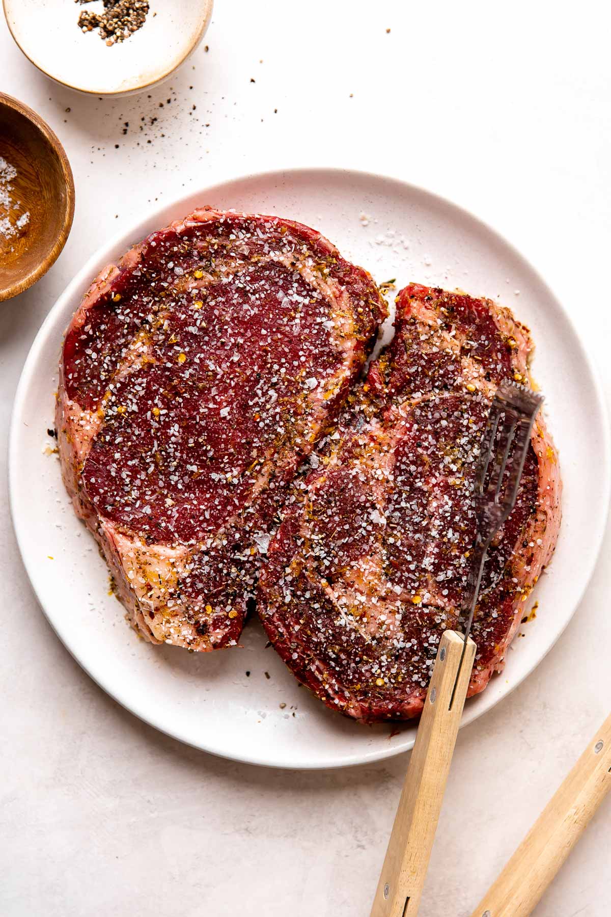 Two steaks marinated with Tuscan style marinade sit atop a white plate that sits atop a creamy white textured surface. The steaks are prepared for grilling by coming up to room temperature and are seasoned with kosher salt and ground black pepper. Two small pinch bowls filled with kosher salt and ground black pepper rest alongside the plate, while a pair of grill tongs rests partially on the steaks.
