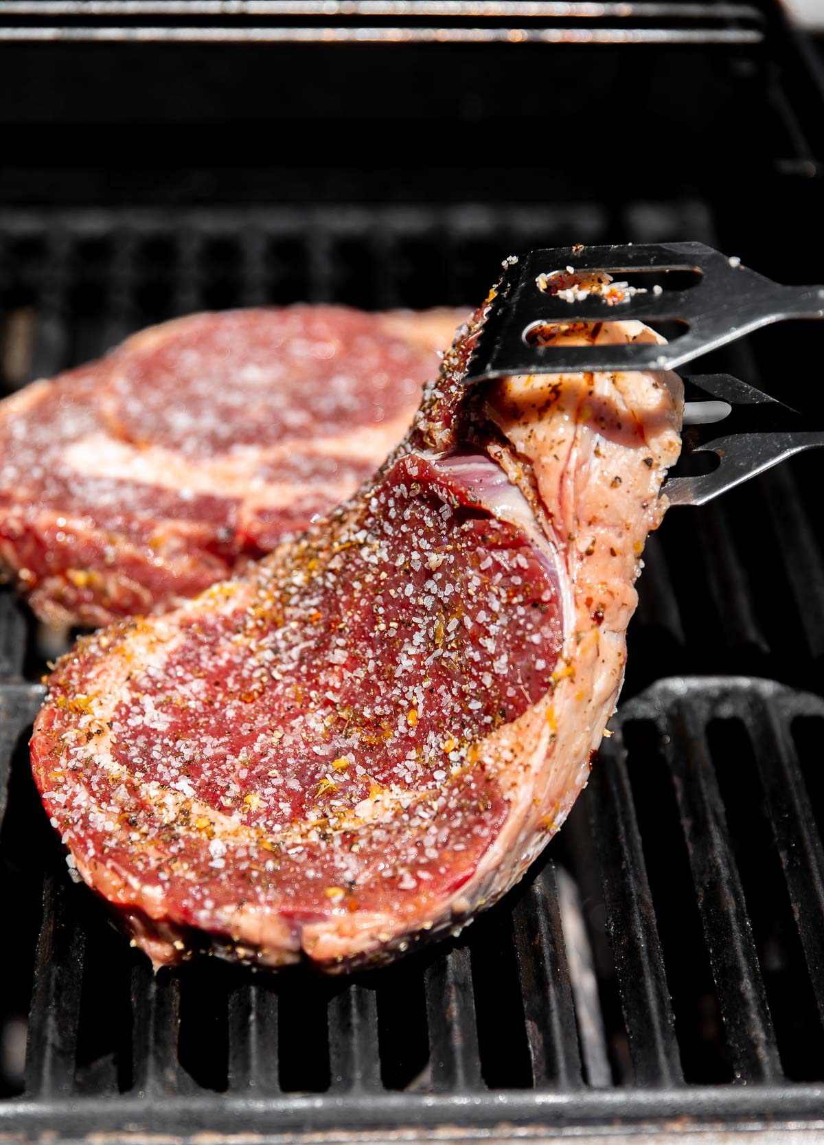 Two Tuscan steaks grill atop gas grill grates. The Tuscan style steak in the foreground is being placed on the grill with a pair of metal grill tongs while the steak in the background already lies flat on the grill.