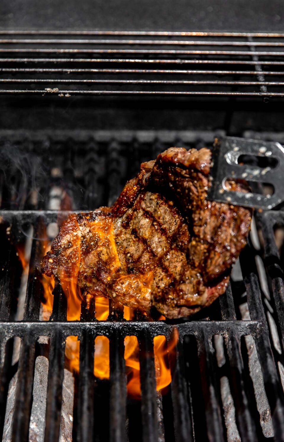 A pair of metal grill tongs works to flip a Tuscan steak over gas grill grates. The steak has visible grill marks and a small flare up of fire can be seen underneath the Tuscan grilled steak.