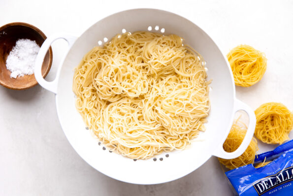 Cooked capellini pasta sits inside of a white colander that sits atop a creamy white textured surface. An open package of dried DeLallo capellini pasta nests rests alongside the white colander while a small wooden pinch bowl filled with kosher salt rests on the other side.