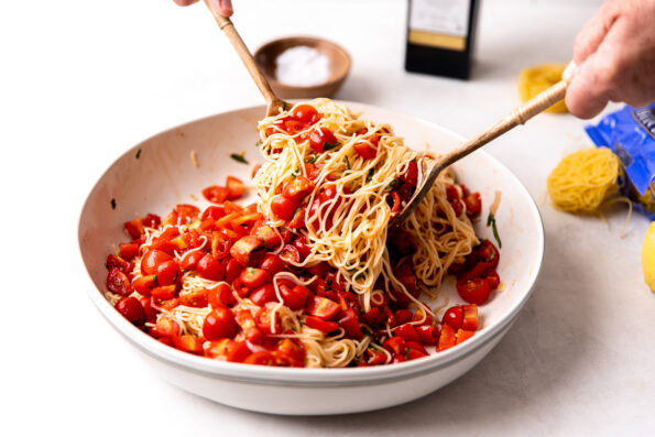 A large white ceramic bowl filled with fresh tomato pasta sits atop a creamy white textured surface. A pair of wooden serving tongs tosses warm capellini pasta with marinated tomatoes and shaved parmesan. A small wooden pinch bowl filled with kosher salt, an open package of DeLallo capellini with dried capellini nests, and a bottle of DeLallo extra virgin olive oil sit behind the bowl in the background.