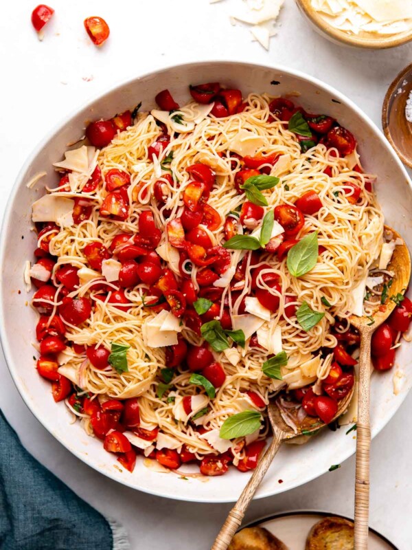 An overhead shot of finished marinated tomato pasta served in a large white ceramic serving bowl with two wooden serving spoons inside. A small wooden pinch bowl filled with shaved parmesan, a small wooden pinch bowl filled with kosher salt, a small ceramic plate of grilled bread, and a blue linen napkin surround the bowl at center.