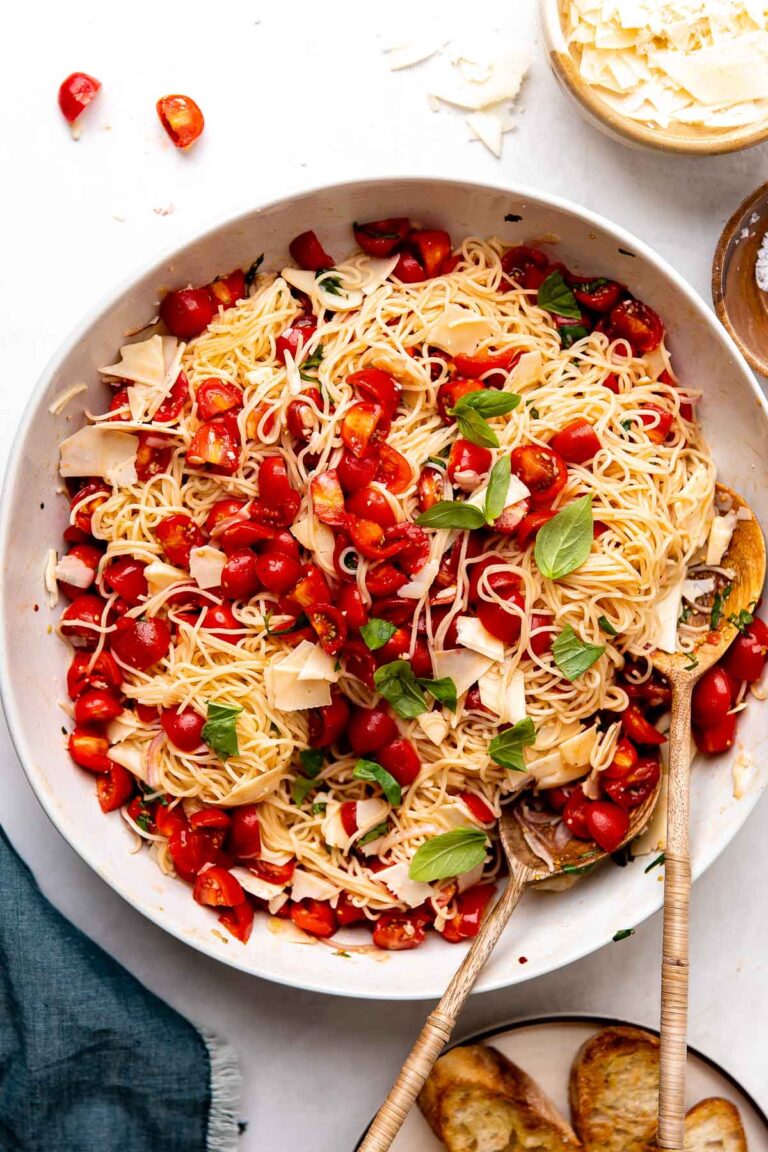 An overhead shot of finished marinated tomato pasta served in a large white ceramic serving bowl with two wooden serving spoons inside. A small wooden pinch bowl filled with shaved parmesan, a small wooden pinch bowl filled with kosher salt, a small ceramic plate of grilled bread, and a blue linen napkin surround the bowl at center.
