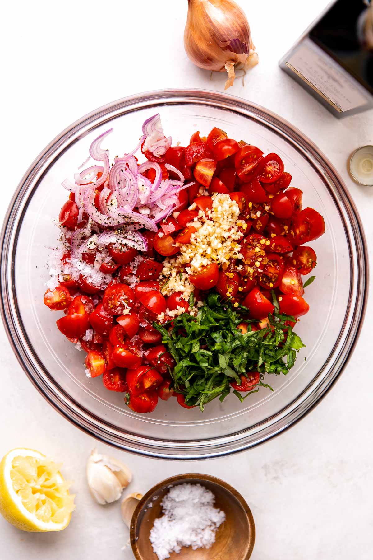 An overhead shot of a clear glass bowl of quartered cherry tomatoes, finely sliced shallot, finely chopped garlic, finely sliced fresh basil, crushed red pepper flakes, and lemon juice sits atop a creamy white textured surface. Extra virgin olive oil has been poured over top of the tomato mixture to allow the tomatoes to marinate. A full shallot, a bottle of DeLallo extra virgin olive oil, a small pinch bowl filled with kosher salt, a couple of cloves of garlic, and a half of a lemon surround the mixing bowl at center.