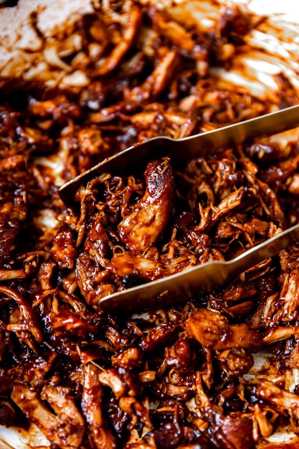 A close up and side angle shot of BBQ shredded chicken inside of a white braiser. A pair of gold tongs rest inside of the pan being used to grab a serving of the BBQ chicken.