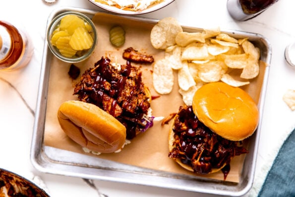 An overhead shot of two shredded BBQ chicken sandwiches arranged atop a parchment lined baking sheet. A small clear glass jar filled with pickles and loose classic potato chips rest alongside the sandwiches. The baking sheet sits atop a white and gray marble surface with a bottle of beer, the beer cap, a small white bowl filled with coleslaw, a bottle of BBQ sauce, a blue linen napkin, and a large white braiser filled with BBQ chicken surrounding the baking sheet at center.