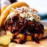 A side angle shot of two BBQ shredded chicken sandwiches arranged atop a parchment lined baking sheet. The sandwiches have been topped with coleslaw and served alongside pickle chips. A small bowl filled with coleslaw rests alongside the baking sheet with a bottle of beer, a bottle of BBQ sauce, and a blue linen napkin shown out of focus in the background.