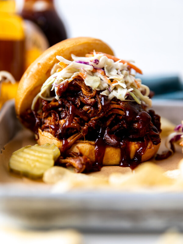 A side angle shot of a single BBQ shredded chicken sandwiches arranged atop a parchment lined baking sheet. The sandwich has been topped with coleslaw and served alongside pickle chips. A bottle of beer, a bottle of BBQ sauce, and a blue linen napkin are shown out of focus in the background.