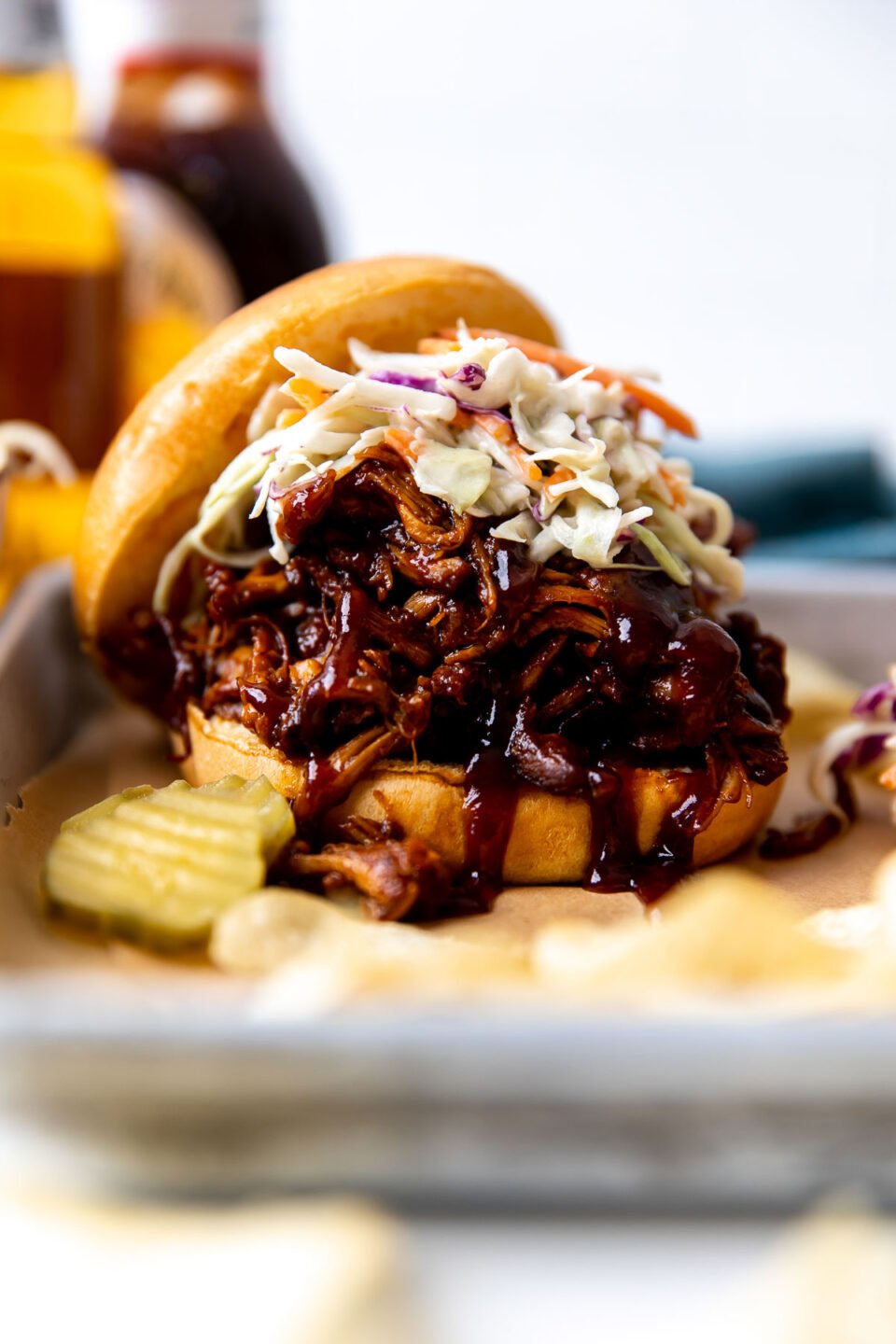 A side angle shot of a single BBQ shredded chicken sandwiches arranged atop a parchment lined baking sheet. The sandwich has been topped with coleslaw and served alongside pickle chips. A bottle of beer, a bottle of BBQ sauce, and a blue linen napkin are shown out of focus in the background.