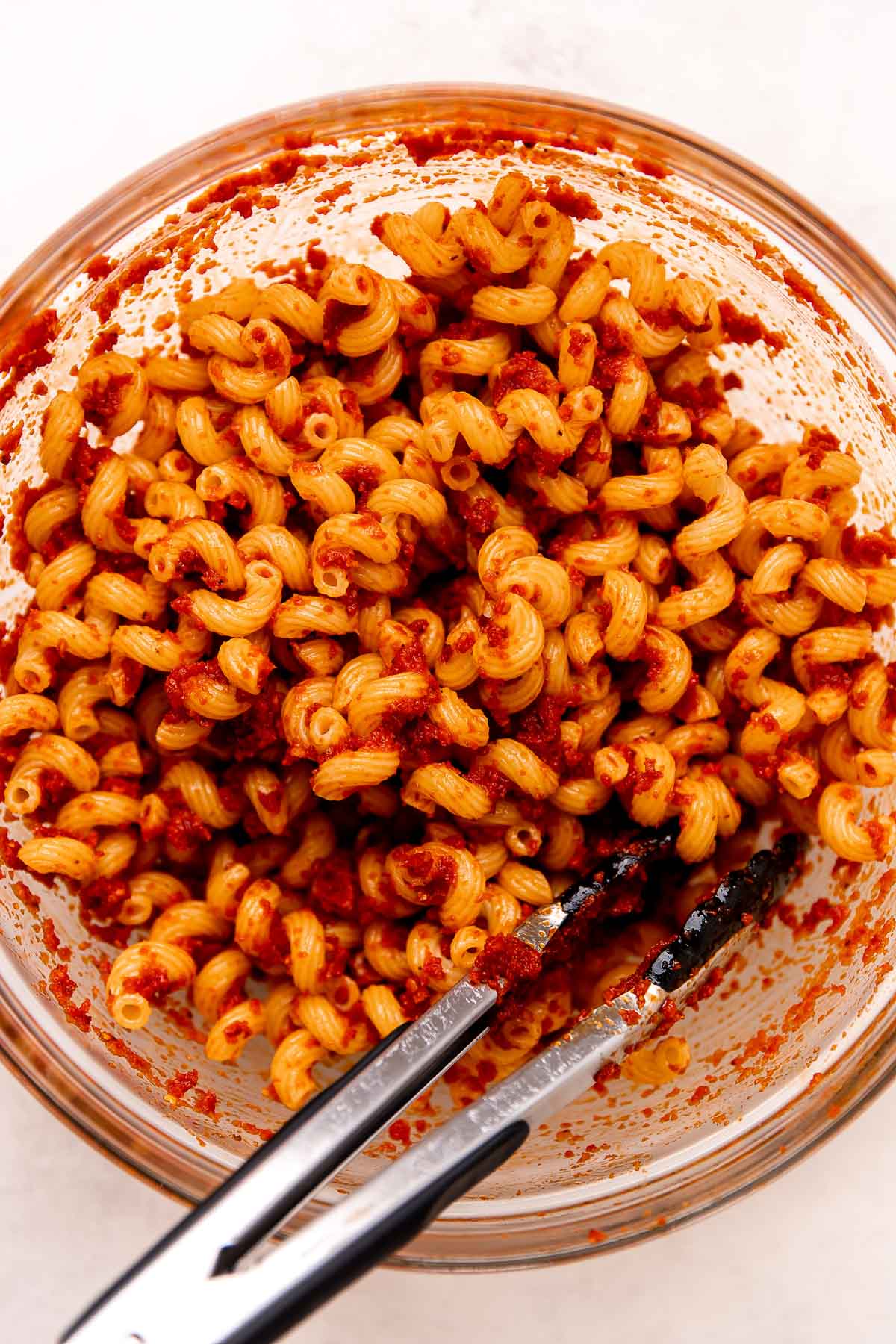Cooked cavatappi pasta tossed with sundried tomato dressing rests inside of a clear glass mixing bowl to make pasta salad with sundried tomatoes. The bowl sits atop a creamy white textured surface. A pair of tongs rests inside of the bowl.