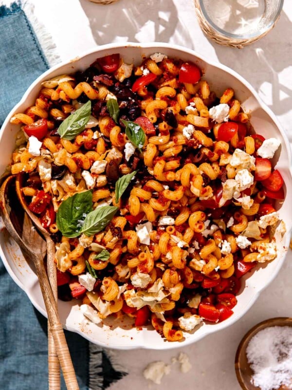 Sundried tomato pasta salad served inside of a large white serving bowl with scalloped edges sits atop a creamy white textured surface with an indigo blue linen napkin placed underneath. Two wooden serving spoons rest inside of the pasta salad and the pasta salad has been garnished with fresh basil. Two drinking glasses and a small wooden pinch bowl filled with kosher salt surround the bowl of pasta salad at center.