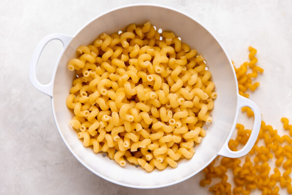 Cooked cavatappi pasta sits inside of a white colander that sits atop a creamy white textured surface. Loose dried cavatappi noodles surround the colander at center.