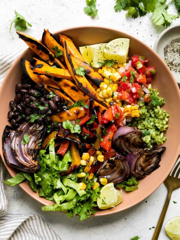 An overhead shot of a grilled vegetable burrito bowl inside of a pink tan ceramic bowl that sits atop a creamy white textured surface. A brown and white linen napkin, a small ceramic pinch bowl filled with ground black pepper, a gold fork, fresh cilantro, and a lime wedge surround the vegetarian burrito bowl at center.