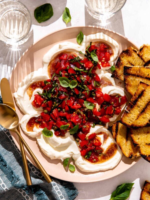 Whipped Ricotta bruschetta served on a peach colored platter garnished with fresh basil and served with grilled bread. The platter sits atop a white textured surface with a blue linen napkin, two glasses of white wine, and a small wooden pinch bowl filled with kosher salt surrounding the platter at center. A gold spoon and knife rest atop the platter for serving.
