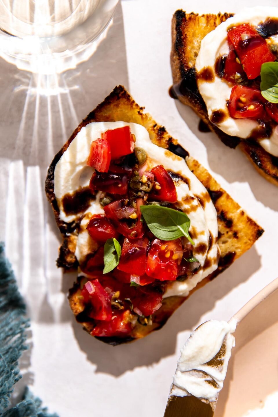 Two pieces of ricotta bruschetta served on grilled bread that sit atop a white textured surface. A blue linen napkin, a peach colored platter with a gold knife used to spread ricotta sitting atop, and a clear glass filled with white wine surround the two pieces of whipped ricotta bruschetta.