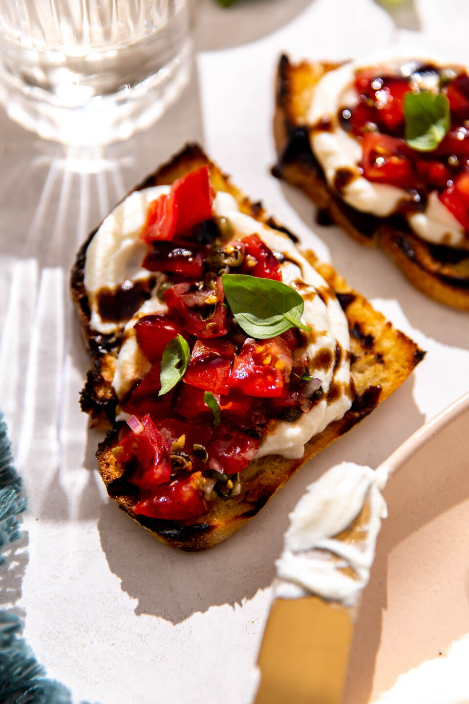 A side angle shot of two pieces of ricotta bruschetta served on grilled bread that sit atop a white textured surface. A blue linen napkin, a peach colored platter with a gold knife used to spread ricotta sitting atop, and a clear glass filled with white wine surround the two pieces of whipped ricotta bruschetta.