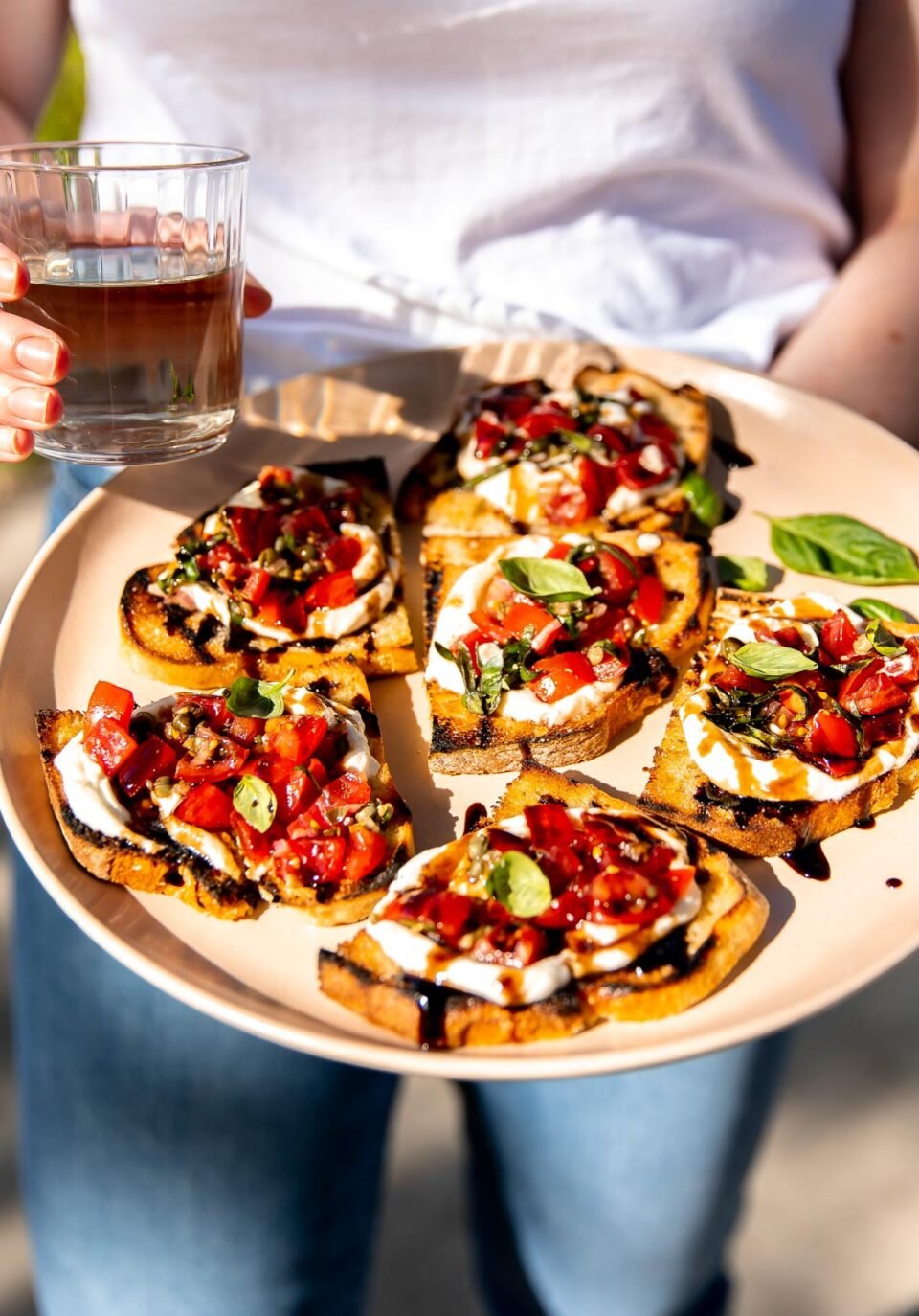 A woman holds a peach colored platter full of pieces of ricotta bruschetta served on grilled bread. The woman holds a clear glass full of white wine and wears jeans and a white tee shirt.