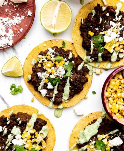 Four assembled black bean tostadas arranged on a gray and white marble surface surrounded by a red ceramic plate topped with cotija cheese, a small ceramic bowl filled with avocado crema with a gold spoon resting inside, a small ceramic bowl filled with kernels of charred sweet corn, and fresh lime wedges surround the bean tostadas.
