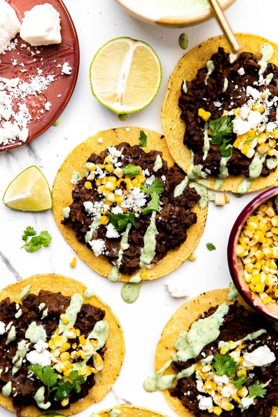 Four assembled black bean tostadas arranged on a gray and white marble surface surrounded by a red ceramic plate topped with cotija cheese, a small ceramic bowl filled with avocado crema with a gold spoon resting inside, a small ceramic bowl filled with kernels of charred sweet corn, and fresh lime wedges surround the bean tostadas.