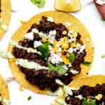 A close up of assembled black bean tostadas surrounded by bowls of toppings and fresh lime wedges.