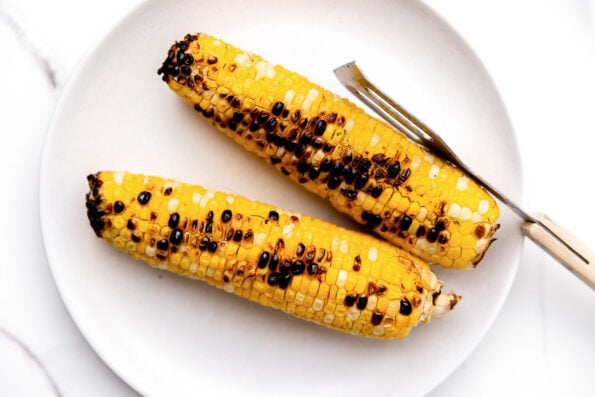 Two ears of charred sweet corn sit atop a white ceramic plate that rests atop a gray and white marble surface. A pair of grilling tongs rests atop the white plate.