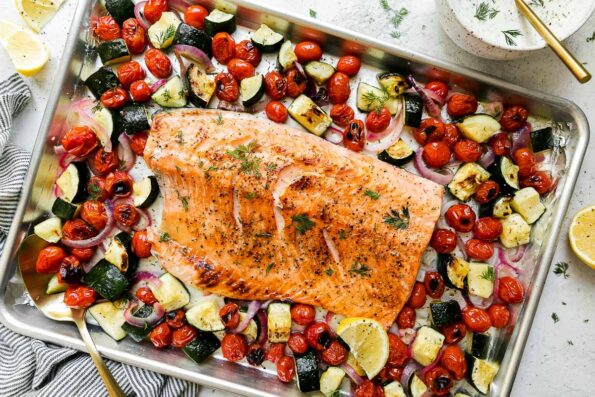 Broiled salmon fillet sits atop an aluminum lined baking sheet pan. The sheet pan salmon is surrounded by summer veggies: burst cherry tomatoes, zucchini, red onion. A small bowl filled with Capered Lemon Dill Sauce, lemon wedges, a blue and white striped linen napkin, and fresh dill surround the sheet pan salmon and veggies at center.