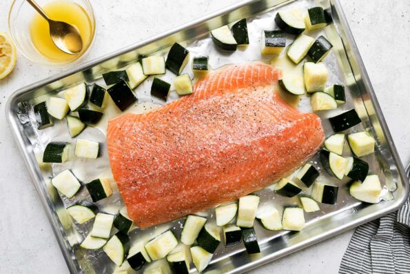 A salmon sheet pan dinner is arranged atop an aluminum baking sheet that sits atop a white textured surface. The salmon fillet is seasoned with kosher salt, ground black pepper, and a lemon honey butter. It is surrounded by 1-inch pieces of zucchini. The baking sheet is surrounded by a small bowl filled with lemon honey butter with a gold spoon in it, a half of a lemon, and a blue and white striped linen napkin.