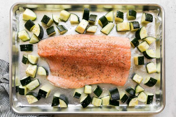A partially baked salmon fillet sits atop an aluminum lined baking sheet surrounded by 1-inch pieces of zucchini. The sheet pan salmon sits atop a white textured surface with a blue and white striped linen napkin underneath the pan.