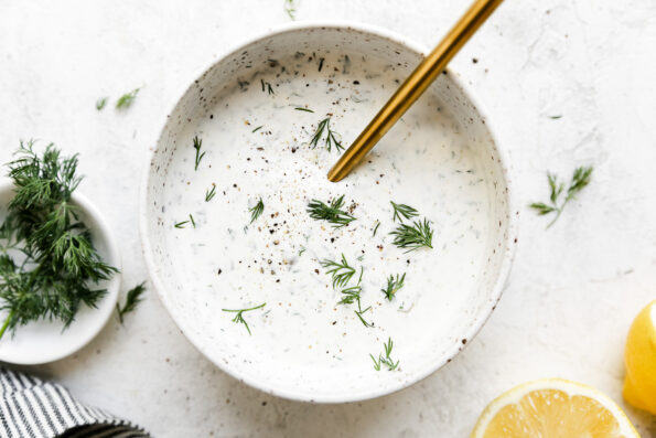 Capered lemon dill sauce fills a white speckled ceramic bowl that sits atop a white textured surface with a gold spoon resting inside of the bowl. The bowl is surrounded by lemon halves, a small pinch bowl filled with fresh dill, and a blue and white striped linen napkin.