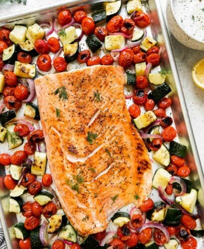 Broiled salmon fillet sits atop an aluminum lined baking sheet pan. The sheet pan salmon is surrounded by summer veggies: burst cherry tomatoes, zucchini, red onion. A small bowl filled with Capered Lemon Dill Sauce, a half of a lemon, a blue and white striped linen napkin, and fresh dill surround the sheet pan salmon and veggies at center.
