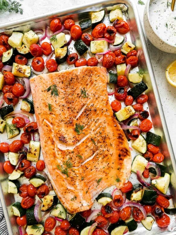 Broiled salmon fillet sits atop an aluminum lined baking sheet pan. The sheet pan salmon is surrounded by summer veggies: burst cherry tomatoes, zucchini, red onion. A small bowl filled with Capered Lemon Dill Sauce, a half of a lemon, a blue and white striped linen napkin, and fresh dill surround the sheet pan salmon and veggies at center.
