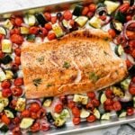 Broiled salmon fillet sits atop an aluminum lined baking sheet pan. The sheet pan salmon is surrounded by summer veggies: burst cherry tomatoes, zucchini, red onion. A half of a lemon, a blue and white striped linen napkin, and fresh dill surround the sheet pan salmon and veggies at center.