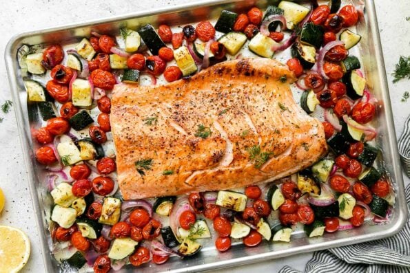 Broiled salmon fillet sits atop an aluminum lined baking sheet pan. The sheet pan salmon is surrounded by summer veggies: burst cherry tomatoes, zucchini, red onion. A half of a lemon, a blue and white striped linen napkin, and fresh dill surround the sheet pan salmon and veggies at center.