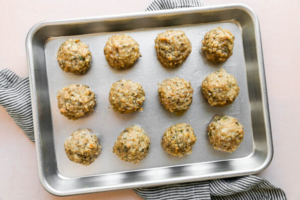 Twelve baked gruyère cheese stuffed meatballs arranged on a small aluminum baking sheet. The baking sheet sits atop a light pink textured surface with a blue and white striped linen napkin placed underneath the pan.