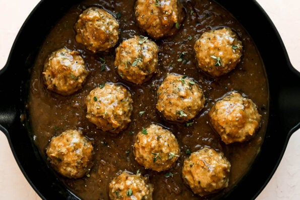 A close up of French onion meatballs nestled inside of a caramelized onion sauce inside of a large gray Stuab cast iron skillet. The skillet sits atop a light pink textured surface and is garnished with ground black pepper and fresh thyme leaves.